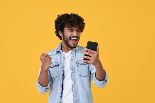 Emocionado joven ganador indio usando teléfono inteligente aislado sobre fondo amarillo