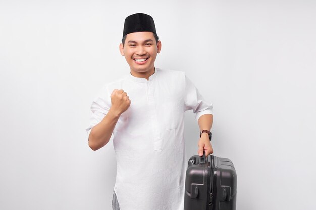 Emocionado joven musulmán asiático de pie mientras sostiene una maleta en la mano celebrando el éxito y listo para la festividad eid Mubarak aislado sobre fondo blanco Concepto de Ramadán y eid Mubarak