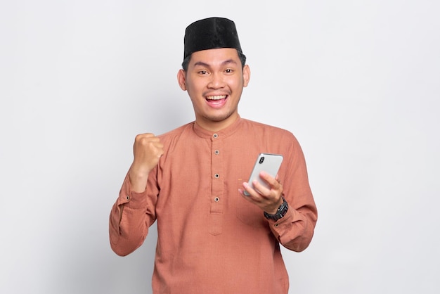 Emocionado joven musulmán asiático usando un teléfono móvil y celebrando el éxito recibiendo buenas noticias aisladas sobre fondo blanco