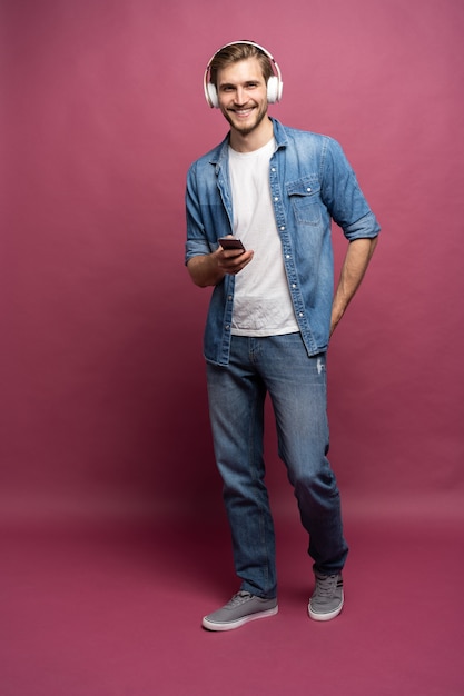 Emocionado joven vestido con camisa de jeans que se encuentran aisladas sobre fondo rosa, escuchando música con auriculares y teléfono móvil