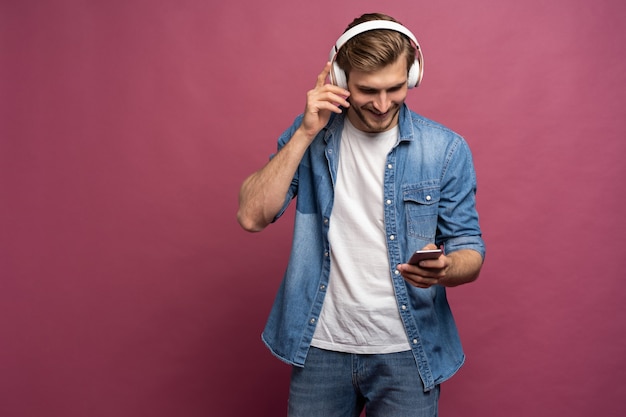 Emocionado joven vestido con camisa de jeans que se encuentran aisladas sobre fondo rosa, escuchando música con auriculares y teléfono móvil