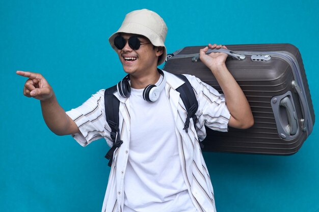 Emocionado joven viajero turista en ropa de verano sombrero sostener maleta punto dedo índice a un lado isol