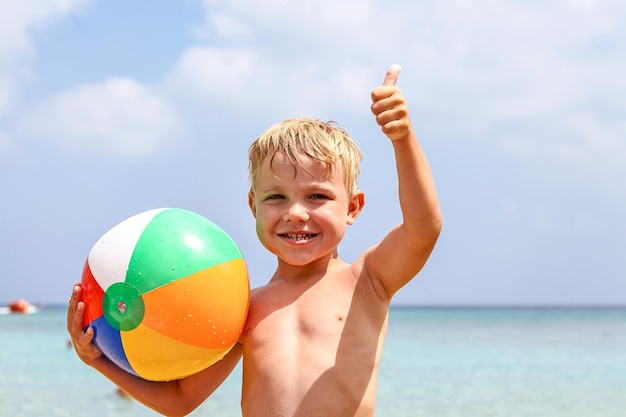 Emocionado niño riéndose sosteniendo una colorida pelota de playa en la playa disfrutando de las vacaciones de verano