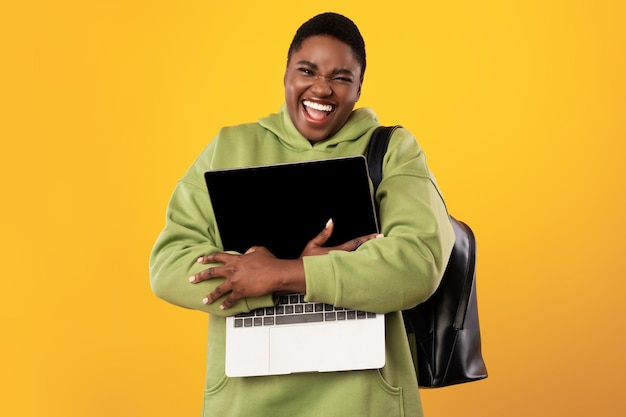 Emocionado PlusSized Black Lady abrazando la computadora portátil sobre fondo amarillo