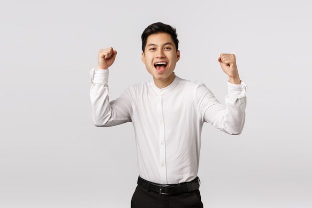 Emocionado, regocijado joven empresario asiático exitoso celebrando la victoria, convertirse en campeón, lograr el objetivo, dar puñetazos y gritar sí como satisfecho, el equipo ganó, regocijarse por la victoria,