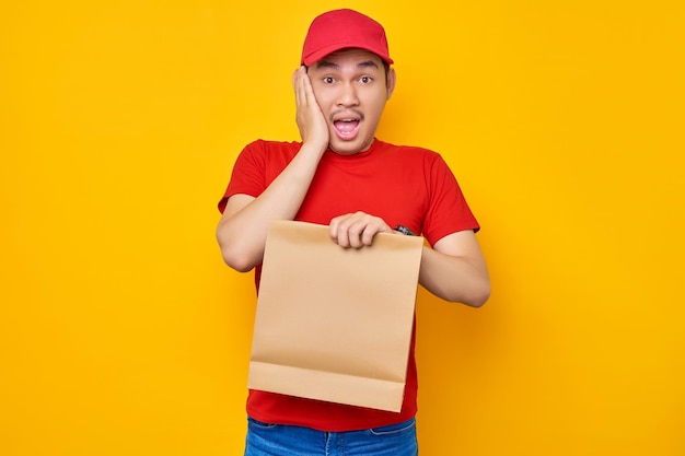 Emocionado repartidor asiático con camiseta roja que trabaja en el servicio de mensajería desde el restaurante de la tienda hasta la oficina en casa con una maqueta de bolsa de comida para llevar de papel artesanal marrón aislada sobre fondo amarillo