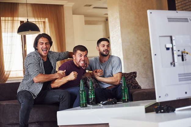 Emocionados tres amigos viendo fútbol en la televisión en casa juntos.