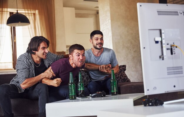 Emocionados tres amigos viendo fútbol en la televisión en casa juntos.