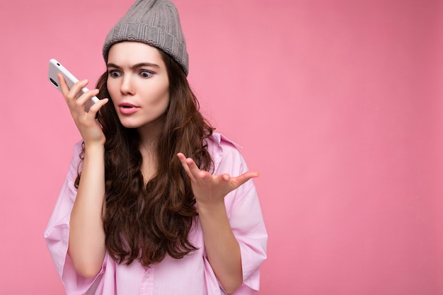 Emocional atractiva joven morena vestida con elegante camisa rosa y sombrero gris aislado