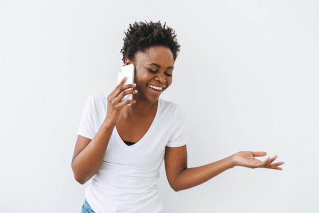 Emocional bela jovem mulher afro-americana em camiseta branca usando telefone celular isolado