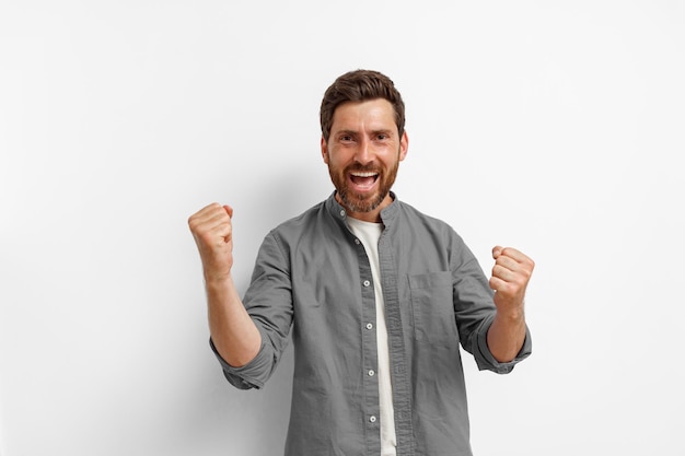 Foto emocional fan masculino sonriente está de pie sobre fondo blanco y gritando con los brazos levantados