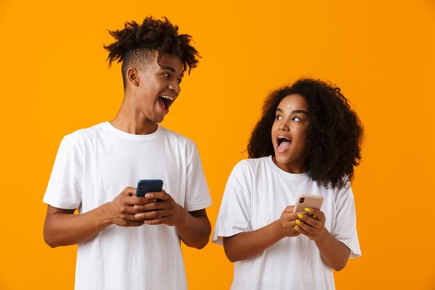 Emocional feliz jovem fofo casal africano isolado sobre o espaço amarelo usando telefones celulares.