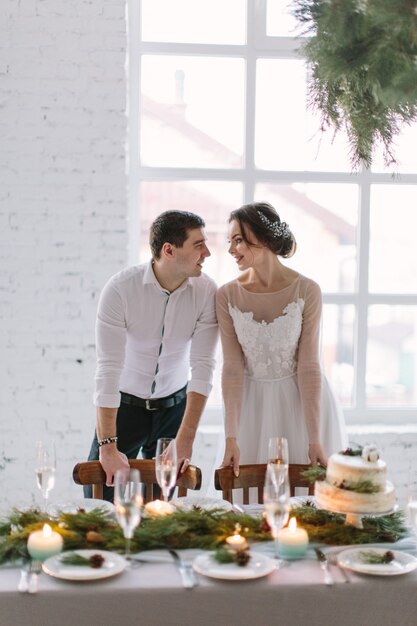 Emocional hermosa pareja de recién casados sonriendo, besándose y abrazándose en la recepción de la boda en el salón blanco con decoraciones de boda