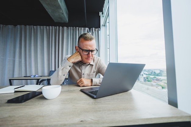 Emocional jovem gerente na mesa de trabalho com laptop em óculos de rosto Um empresário alegre está trabalhando em um escritório moderno com grandes janelas