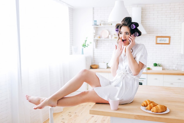 Emocional joven descuidada sentarse en la mesa y hablar por teléfono. Ama de casa sorprendida en la cocina. Vistiendo bata blanca. La vida sin trabajo. Elegante mujer con rulos en el cabello.