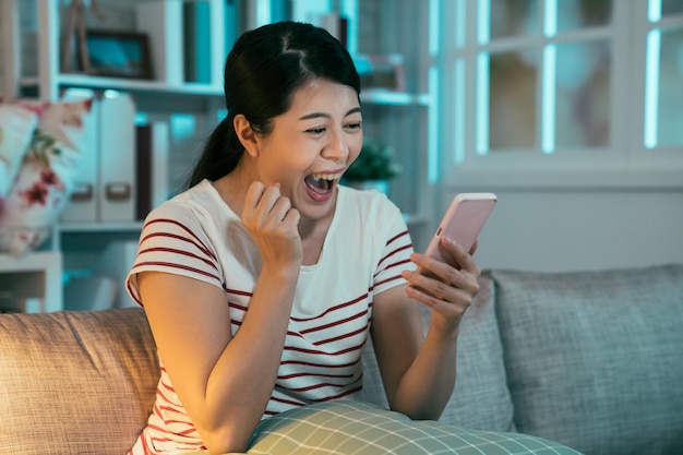 Emocional mujer asiática que se siente emocionada por recibir buenas noticias sosteniendo un teléfono inteligente sentado en un sofá en casa por la noche. niña feliz gritando sí feliz por el éxito en la competencia en línea verificar el teléfono de correo