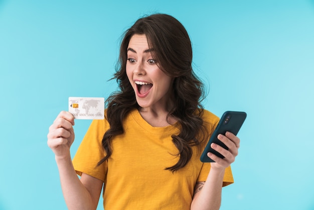 Emocional sorprendido sorprendido joven bonita a mujer posando aislada sobre pared azul mediante teléfono móvil con tarjeta de crédito.