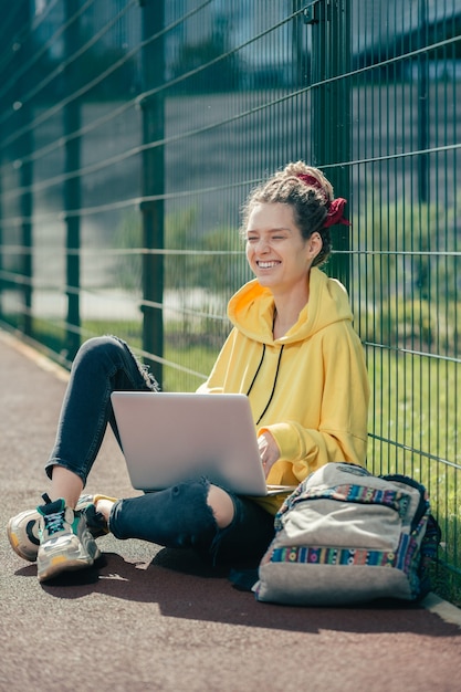 Emocionalmente positiva, jovem segurando um laptop moderno e rindo com os olhos fechados enquanto está no campo de esportes