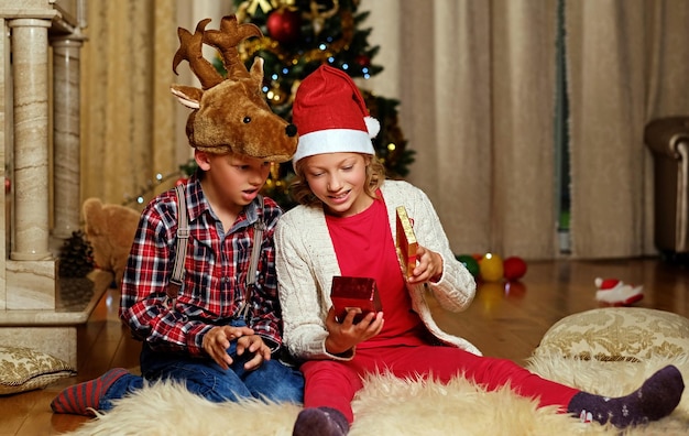 Emocionante chico lindo con sombrero de ciervo de Navidad y chica feliz sostiene una caja de regalo en una habitación decorada con Navidad.