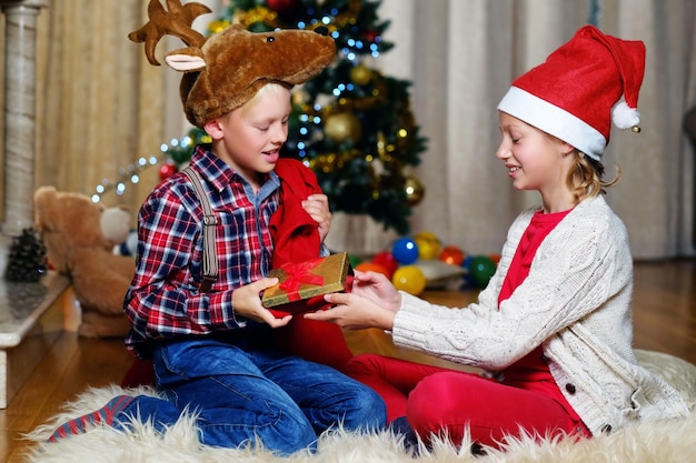 Emocionante menino bonitinho com chapéu de veado de natal e garota feliz segura caixa de presente na sala decorada de natal.