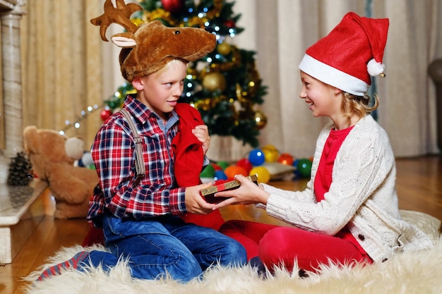 Emocionante menino bonitinho com chapéu de veado de Natal e garota feliz segura caixa de presente na sala decorada de Natal.