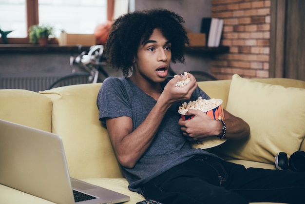 Emocionante programa de TV. Jovem africano assistindo TV e mantendo a boca aberta enquanto está sentado com um balde de pipoca no sofá de casa