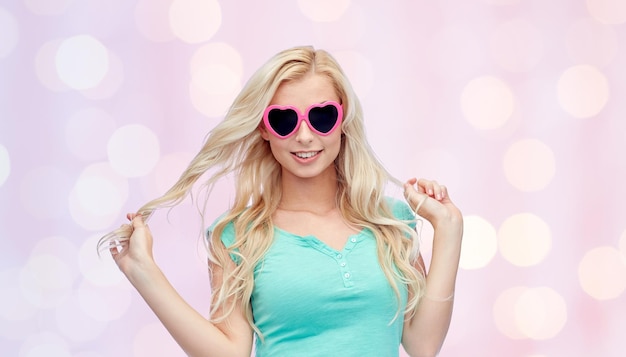 emociones, expresiones, concepto de verano y personas - mujer joven sonriente o adolescente con gafas de sol en forma de corazón sosteniendo su cabello sobre fondo de luces de vacaciones rosadas