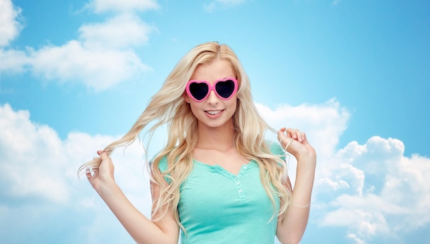 emociones, expresiones, verano y concepto de la gente - mujer joven sonriente o adolescente con gafas de sol en forma de corazón sosteniendo su cabello sobre el cielo azul y el fondo de las nubes