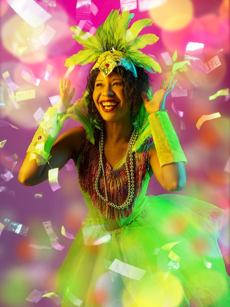 Emociones Hermosa mujer joven en carnaval, elegante disfraz de mascarada con plumas sobre fondo degradado en neón, confeti volador. Celebración de fiestas, baile, moda. Tiempo festivo, fiesta.