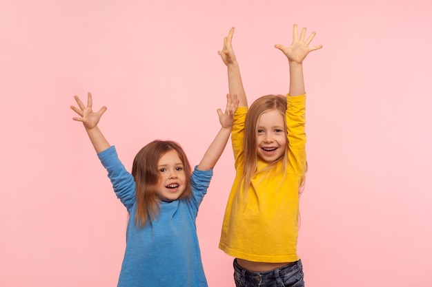 Emociones de los niños ganadores felices Dos niñas entusiastas y enérgicas de pie con las manos levantadas y gritando de alegría celebrando la victoria en un estudio interior aislado en un fondo rosa