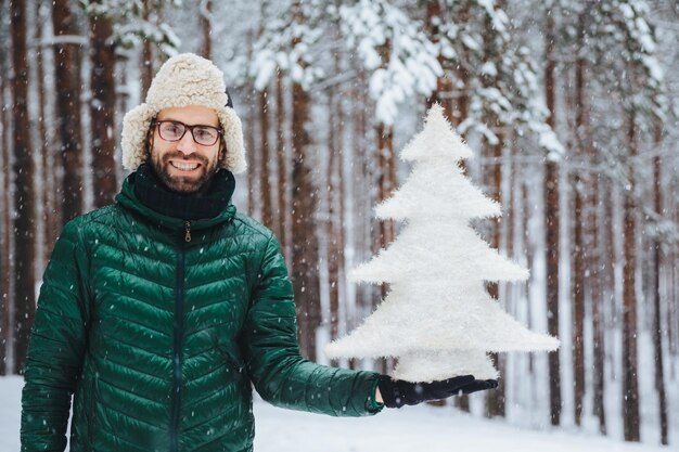 Emoções de pessoas clima nevado e conceito de temporada Feliz e alegre jovem homem com barba por fazer vestido com roupas quentes segura abetos contra o fundo das árvores desfruta de belas paisagens naturais