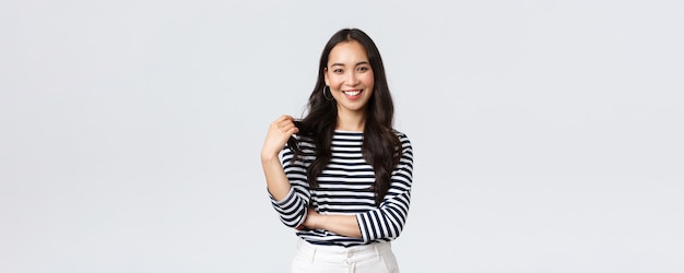 Emoções de pessoas de estilo de vida e conceito casual Encantadora garota coreana sorridente em camisa listrada tocando o fio de cabelo e sorrindo alegremente câmera falando com um amigo fundo branco