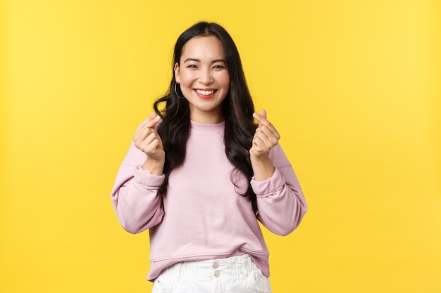 Emoções de pessoas, estilo de vida e conceito de moda. Menina asiática de kawaii com um lindo sorriso, mostrando o gesto de corações coreanos, enviando vibrações positivas e alegres, em pé fundo de anúncio amarelo.