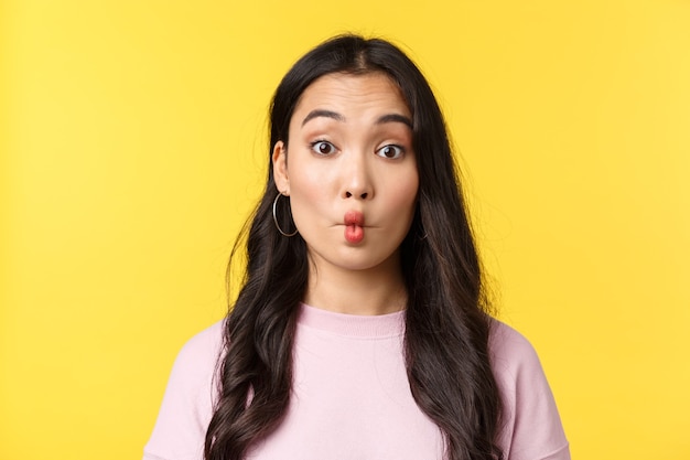 Emoções de pessoas, lazer do estilo de vida e conceito de beleza. Menina asiática boba e engraçada fazendo lábios de peixe e olhando a câmera indecisa, comendo algo azedo, fundo amarelo de pé.