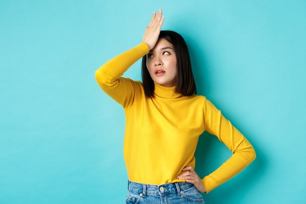 Emoções e conceito de estilo de vida. Menina asiática irritada revira os olhos e a palma do rosto, em pé incomodado em uma camisola amarela contra um fundo azul.