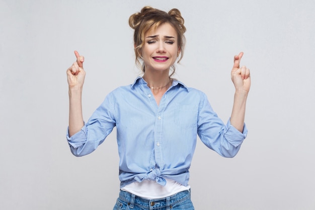 Emoções e sentimentos humanos. menina adolescente supersticiosa com cabelos loiros e rosto bonito cruzando os dedos para dar boa sorte, esperando que seus desejos se tornem realidade, tendo olhar feliz animado.