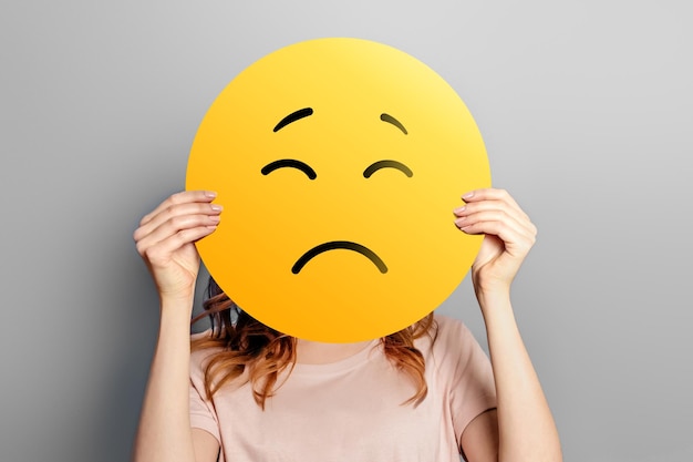 Emoji triste Garota segura um emoticon amarelo com rosto triste isolado em um emoji infeliz de fundo cinza