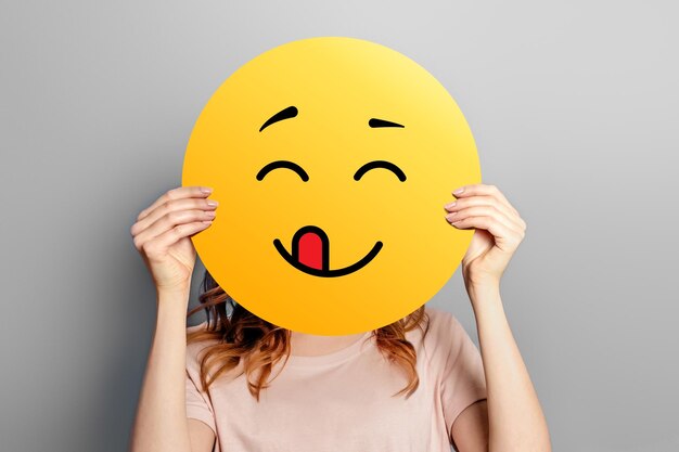 Foto emoticon lecken mädchen hält einen gelben smiley mit zunge isoliert auf grauem hintergrund