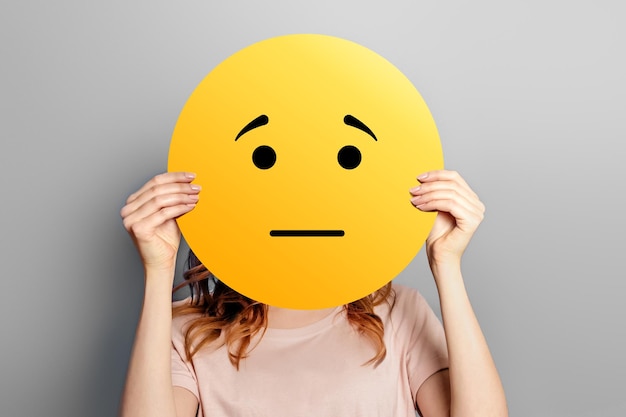 Emoticon ofendido Garota segura um smiley amarelo com rosto triste isolado em um fundo cinza