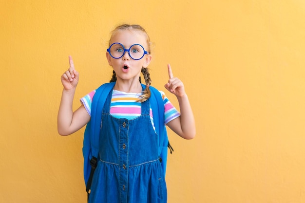 Emotional aufgeregtes Überraschungsgesicht Schulmädchen Kind in Brille in blauer Schuluniform mit offenem Mund der Rucksackschultasche zeigen die Finger bis zum leeren Kopierraum aufmerksam isoliert auf gelbem Hintergrund