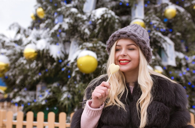 Emotionale blonde Frau in grauer Strickmütze, die köstliche Pralinen gegen den Weihnachtsbaum in der Stadt hält