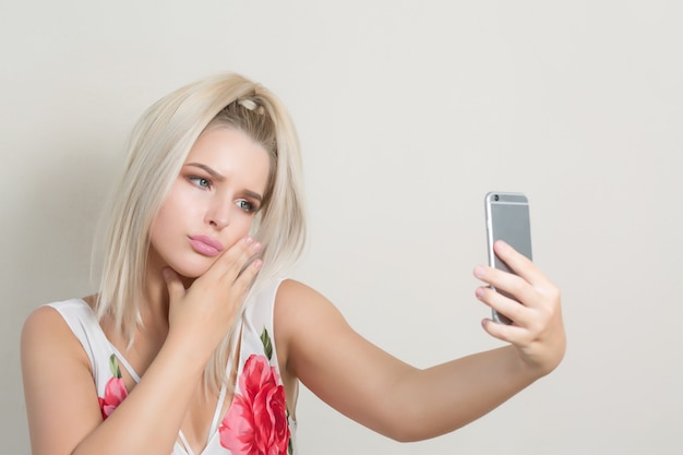 Emotionale blonde Frau macht Selfie auf dem Handy vor einem grauen Hintergrund