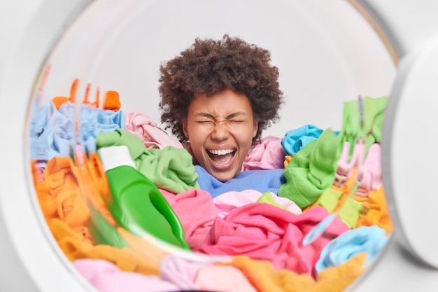 Emotionale Frau mit lockigem Haar steckt den Kopf in einen Haufen bunter Wäsche mit einer Flasche flüssigem Pulver, die damit beschäftigt ist, Posen aus der Waschmaschine zu waschen