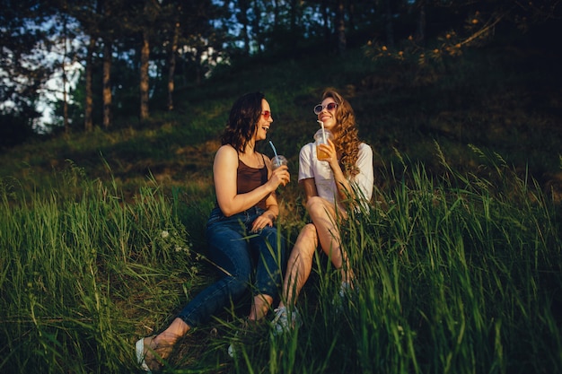 Emotionale Freundinnen werden fröhlich, setzen sich ins Gras, trinken einen Cocktail mit Sonnenbrille in weißem und schwarzem Hemd, bei Sonnenuntergang, positiver Gesichtsausdruck, im Freien