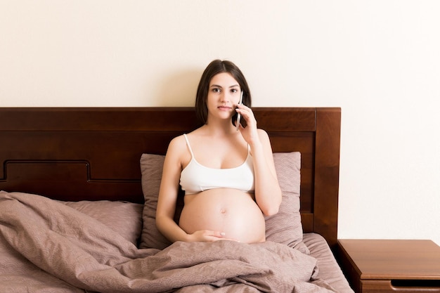 Emotionale schwangere Frau, die zu Hause im Bett mit dem Handy telefoniert