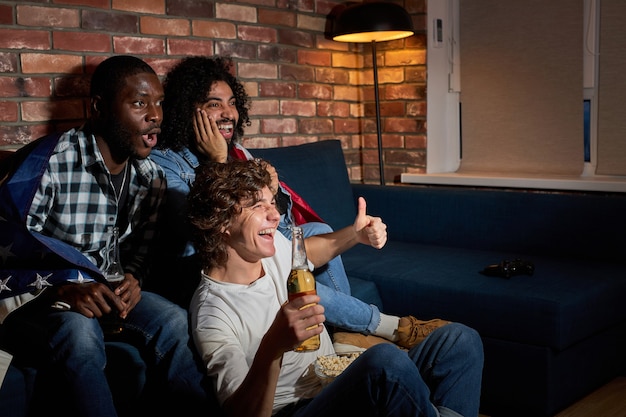 Emotionale Studenten, die zu Hause Sportmatches auf dem Sofa sitzen. Fans jubeln für die amerikanische Lieblingsnationalmannschaft und trinken Bier. Konzept der Emotionen, Unterstützung.