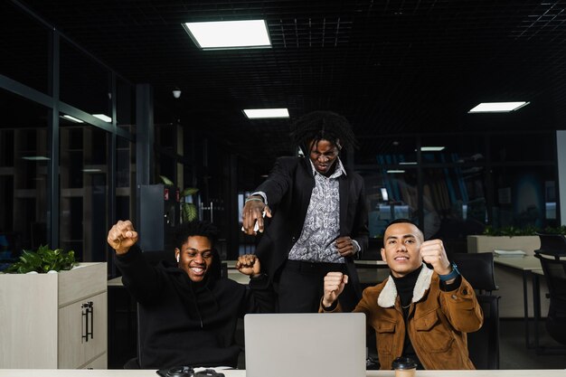 Emotionale und fröhliche Schwarzafrikaner und Asiaten arbeiten zusammen und freuen sich über das Ende des Online-Projekts. Multiethnische Kollegen, die am Laptop arbeiten, heben ihre Hände und schreien laut