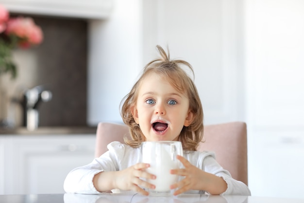 Emotionaler Ausdruck Kindermädchen offener Mund mit Schnurrbart Milch auf den Lippen, die ein Glas Milch halten. Weißlichtküche mit Blumenhintergrund. Milch, Milchprodukte, Kalzium für eine gute Kindergesundheit