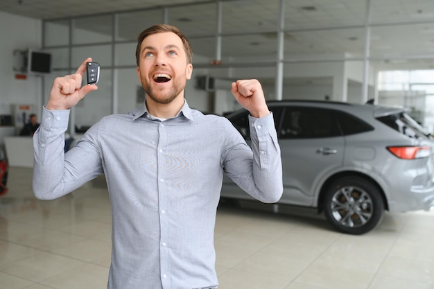 Emotionaler gutaussehender Kerl, der die Hand hochhebt und in die Kamera lächelt Reicher Mann kauft Auto im Luxus-Händlersalon