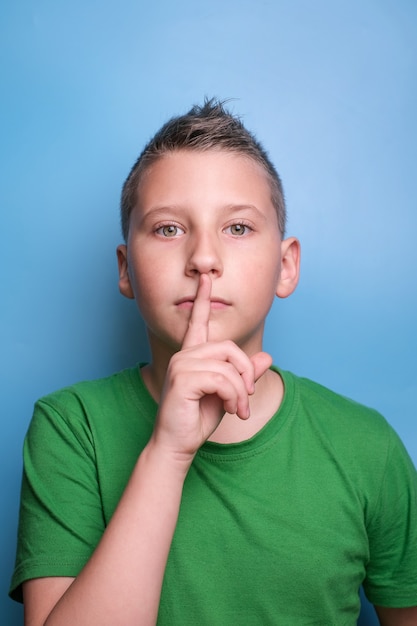 Emotionaler Junge, der den Mund mit dem Finger bedeckt, der zum Schweigen gebracht wird, Porträt auf blauem Hintergrund